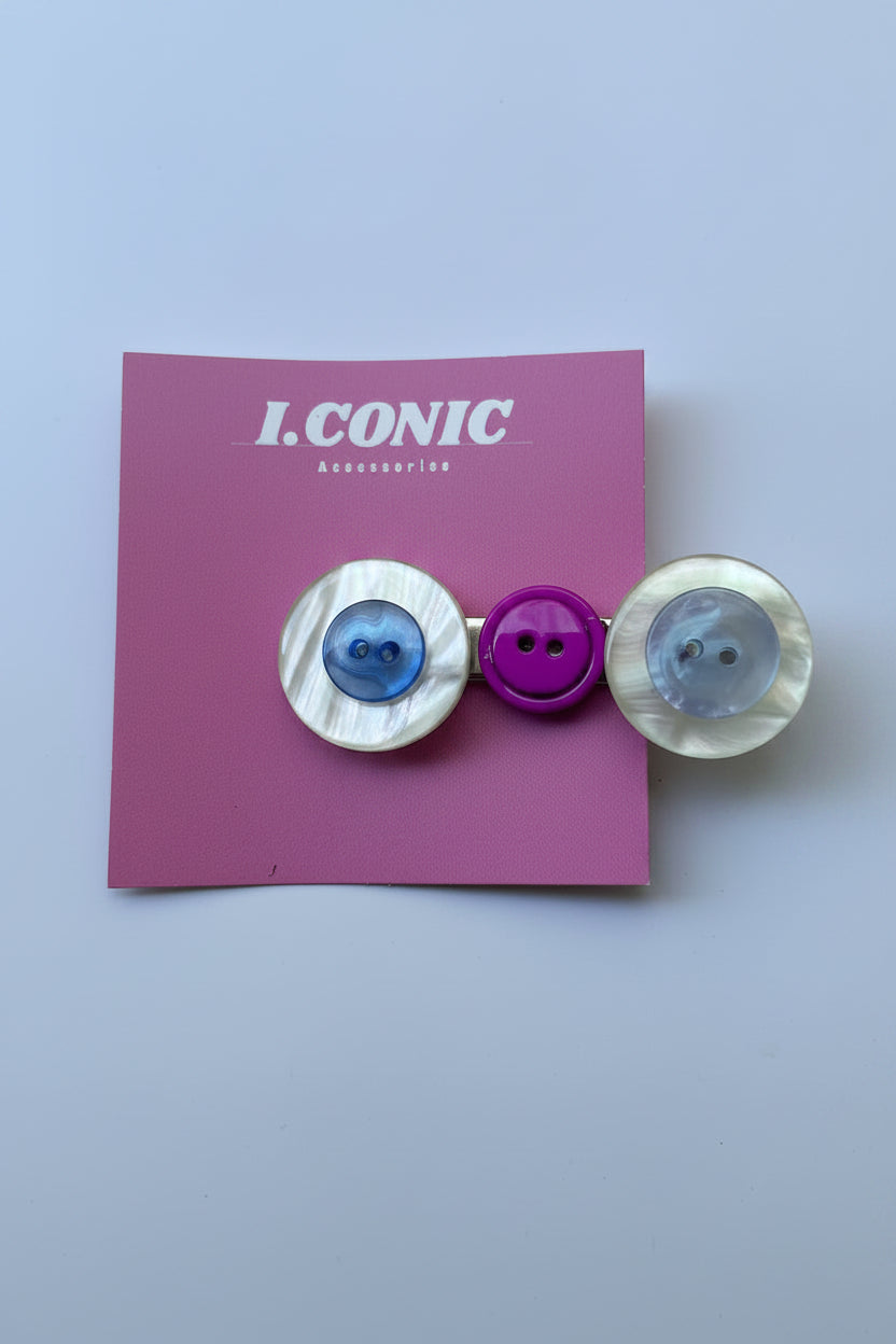 Three decorative buttons attached to a pink card labeled 'ICONIC' against a light gray background. The set includes two round mother-of-pearl buttons (one blue, one clear) and one smaller purple button.